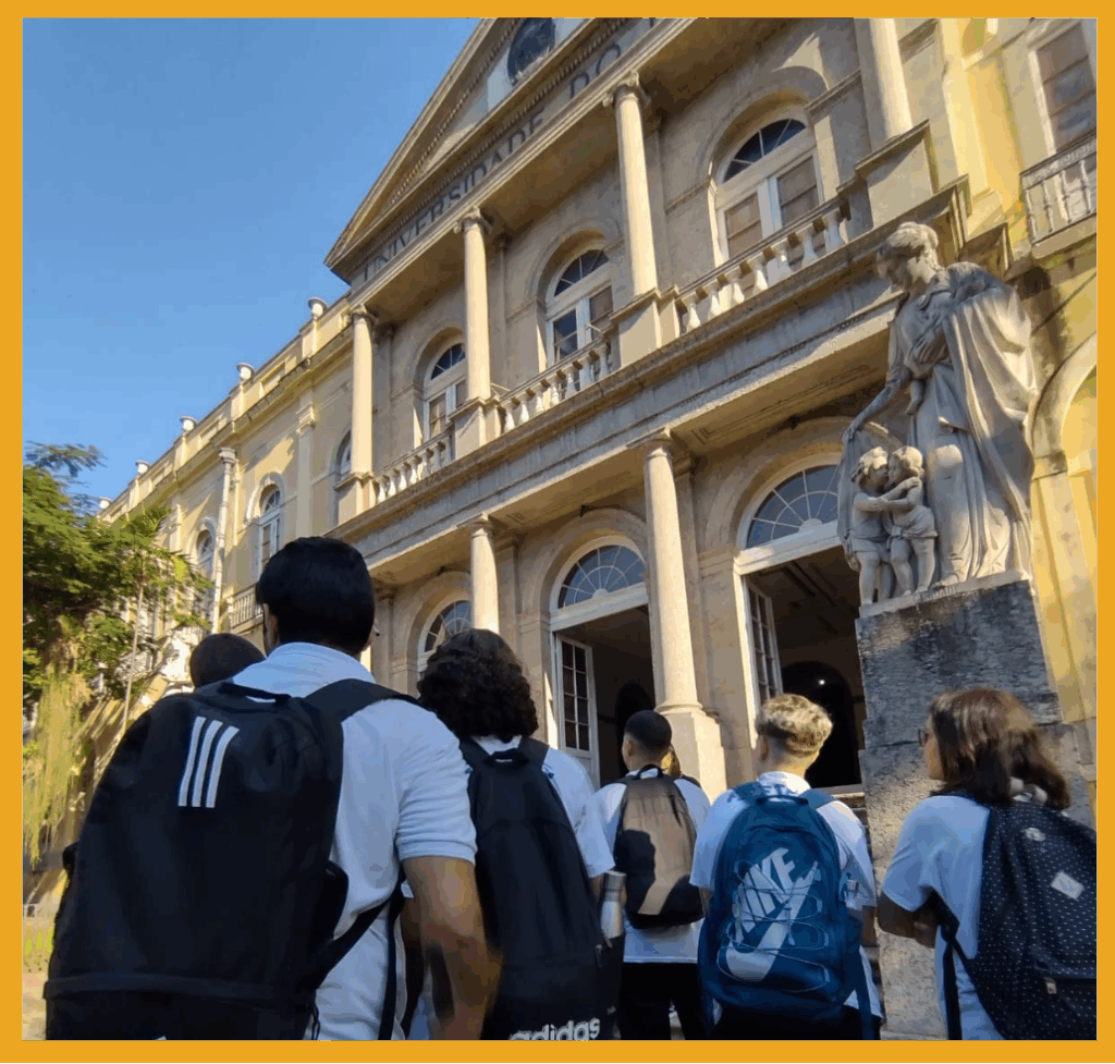 Fotografia dentro de uma borda amarela. Nela, estudantes de costas, observam a fachada do Palácio universitário da UFRJ. Fim da audiodescrição.