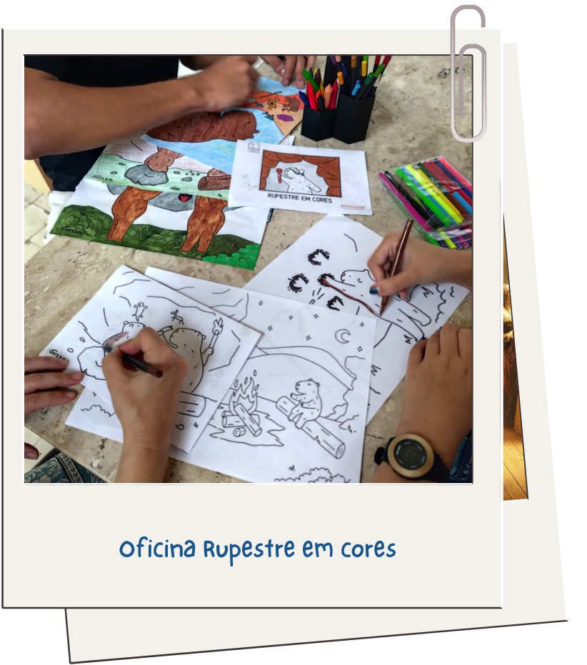 Fotografia de mãos de participantes colorindo desenhos em papel com canetinhas e giz de cera. Os desenhos trazem figuras de estilo rupestre e cenas com animais e fogueira. Sobre a mesa também há um pote com lápis coloridos e canetas. Na parte inferior da imagem está escrito: “Oficina Rupestre em cores”. Fim da audiodescrição.